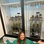 Kate Lucas poses by two racks of sediment cores in a large incubator. on July 2, 2024